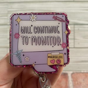 May include: A badge reel with a purple background and the text "WILL CONTINUE TO MONITOR". The badge has a pink glitter border and includes medical symbols, flowers, and the hashtag #NURSELIFE.