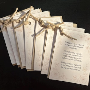 May include: A stack of rectangular off-white paper tags tied together with tan jute twine. Each tag features a light brown floral border and printed text: The paper I'm attached to is Plantable Wildflower Seed Paper! This plantable paper is made from recycled unbleached paper waste and embedded with seeds. When the paper is planted in soil, the seeds grow and the paper composts away.