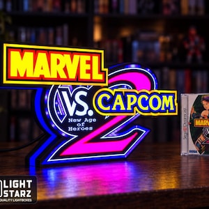May include: A glowing LED sign featuring the Marvel vs. Capcom 2 New Age of Heroes logo with yellow, blue, and pink illumination. Beside the sign sits a Sega Dreamcast video game case for Marvel vs. Capcom 2, displaying character artwork.