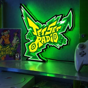 May include: A wall-mounted LED light sign featuring the Jet Set Radio logo in yellow and green with a black outline. The sign emits a bright green glow from behind its edges, displaying the stylized text Jet Set Radio in a graffiti-inspired font.