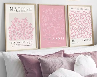 May include: Three framed art prints with beige wooden frames. The prints feature pink and white abstract designs and text, including "MATISSE" and "PICASSO." The prints are displayed above a bed with pink and white pillows.