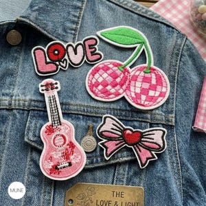 May include: A denim jacket decorated with embroidered patches. Patches include the word "LOVE", cherries, a ukulele, and a bow with a heart. The patches are mainly pink and red with white and black accents. A small metal button and a tag with text are also visible.