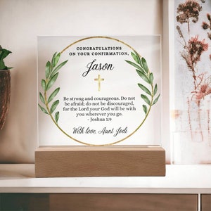Puede incluir: Una placa de acrílico transparente con un marco dorado y hojas verdes alrededor de los bordes. La placa tiene una cruz dorada y el texto "CONGRATULATIONS ON YOUR CONFIRMATION, Jason" y un versículo bíblico de Josué 1:9. El texto "With love, Aunt Jodi" está en la parte inferior de la placa.
