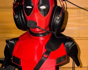 May include: A red and black Deadpool bust with a headset and microphone. The bust has white eyes and a black base with the Deadpool logo. The headset is black with red accents.