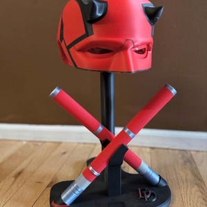 May include: A red and black helmet with horns, resembling a superhero's mask, is displayed on a black stand. Two red batons with silver accents are crossed in front. The base is black, and the background is a brown wall and wooden floor.