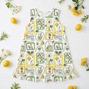 May include: White sleeveless dress with a repeating pattern of cartoon ducks, lemons, lemonade, and bees. The dress has a ruffled hem and a scoop neckline. The design includes the word "FRESH" in a circular frame. Yellow lemons and white flowers are scattered around the dress.