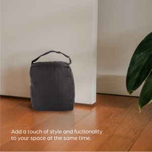 May include: A dark gray fabric doorstop with a loop handle. The doorstop is sitting on a wooden floor.