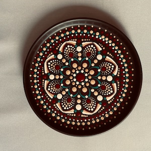 Może przedstawiać: Okrągły, czarny talerz z namalowanym wzorem mandali. Mandala przedstawia centralny kwiat z płatkami w odcieniach czerwieni, bieli i złota, otoczony koncentrycznymi okręgami kropek w różnych kolorach.