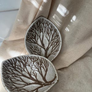 Puede incluir: Dos cuencos de cerámica con forma de hoja y diseño de ramas de árbol marrón. Los cuencos son blancos con una textura moteada y están sobre una tela beige. El diseño de ramas de árbol cubre el interior de cada cuenco.