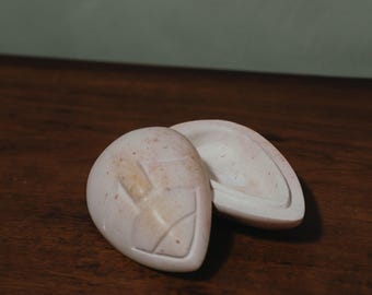May include: Two off-white, egg-shaped stone objects with a carved design on the front, one open to reveal the interior. The objects are on a dark wood surface.