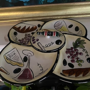 May include: A multi-sectioned serving dish with a wine theme. The dish is cream-colored with black trim and features illustrations of grapes, cheese, wine glasses, and bread. A small, round lid with a bottle stopper is in the center.