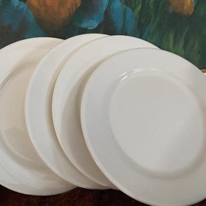 May include: Four white ceramic plates are stacked. Each plate has a broad rim and a gently indented centre. The plates are arranged in a fan shape, with the edges of the plates visible. The plates are clean and appear unused.