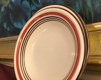 May include: White ceramic bowl with a red, black, and brown striped rim. The bowl has a simple, classic design, suitable for serving soups, salads, or other dishes. The bowl is set against a dark background.