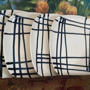 May include: A set of four square ceramic plates with rounded corners, featuring an off-white base color and a hand-painted black grid pattern consisting of intersecting vertical and horizontal lines of varying thickness across the surface.