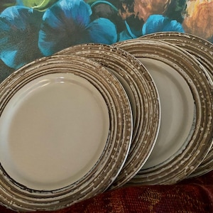 May include: A set of four ceramic plates. Each plate has a cream-colored center and a brown, textured rim with white speckles. The plates are stacked slightly, showcasing their design. The background features a floral painting.