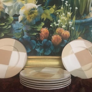 May include: A set of six decorative plates with a geometric design. Each plate has a square pattern in beige, white, and tan, with a gold rim. The plates are arranged on a dark surface, with a floral painting in the background.