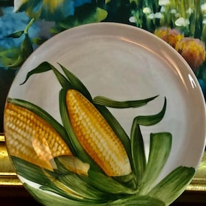 May include: A white ceramic plate with a hand-painted design of two ears of corn. The corn is yellow with green husks and leaves. The plate has a gold rim and is set against a blurred background.