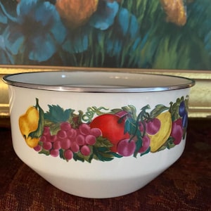 Puede incluir: Un cuenco de esmalte blanco con borde plateado, decorado con una colorida guirnalda de frutas. La guirnalda presenta peras amarillas, tomates rojos, uvas moradas y otras frutas. El cuenco es probablemente para uso en la cocina.