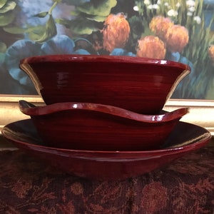 May include: A set of three nesting bowls in a deep burgundy red color. The bowls are made of ceramic and have a glossy finish. The bowls are stacked on top of each other, with the largest bowl on the bottom and the smallest bowl on top.