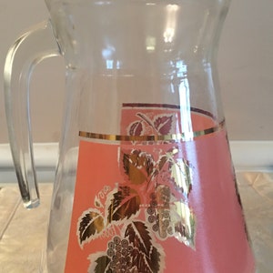 May include: Clear glass pitcher with a handle. The pitcher features a pink panel with gold grape and leaf designs. A gold band accents the top and bottom of the pink panel. The pitcher is likely vintage.