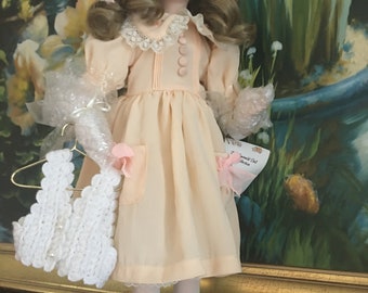 May include: A porcelain doll with blonde hair and blue eyes, wearing a peach-colored dress with a lace collar and a matching bow. The doll holds a white knitted vest and a small bouquet of flowers. The doll is standing on a gray stand.