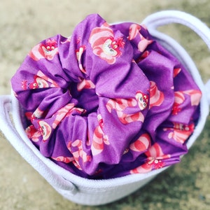May include: Purple scrunchie with a repeating pattern of a cartoon cat with pink and white stripes. The scrunchie is in a white basket.
