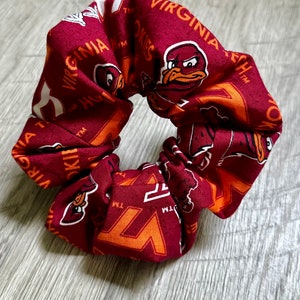 May include: A maroon and orange scrunchie with the Virginia Tech Hokies logo and mascot. The scrunchie is made of fabric and has a white background with the text "Virginia Tech" and "Hokies" in white.