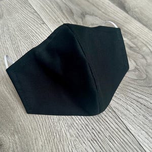 May include: A black face mask with white elastic ear loops. The mask is made of a soft, breathable fabric.