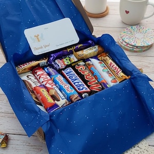 May include: An open cardboard gift box filled with various chocolate candy bars, nestled on blue tissue paper. A white card with text is visible. A small succulent plant and a mug are in the background.
