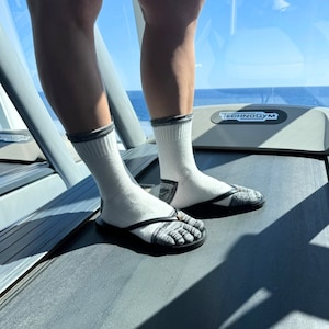 May include: White socks with gray trim and toe-separating flip-flops are worn on a treadmill. The flip-flops have a black sole and a gray and white patterned strap. The treadmill is gray and has the word "TECHNOGYM" printed on it.