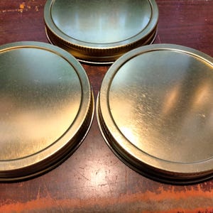 Puede incluir: Tres tapas metálicas doradas para tarros están dispuestas sobre una superficie de madera. Las tapas son redondas con un borde ligeramente elevado y una superficie brillante y reflectante. Las tapas son probablemente para enlatar o conservar alimentos.