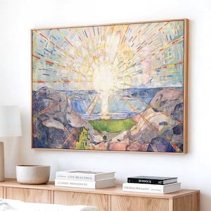 Op de afbeelding: Een rechthoekig ingelijst schilderij met een zonsopgang boven een rotsachtig landschap in blauwe, gele en groene tinten. Het kunstwerk toont stralende lichtbundels boven een horizonlijn, aan een witte muur boven een houten dressoir.