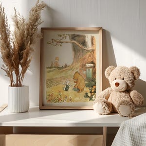 May include: A framed illustration of Winnie the Pooh and Piglet walking in a forest. The illustration is in a light brown frame and is sitting on a white shelf. A brown teddy bear is sitting on the shelf to the right of the frame.