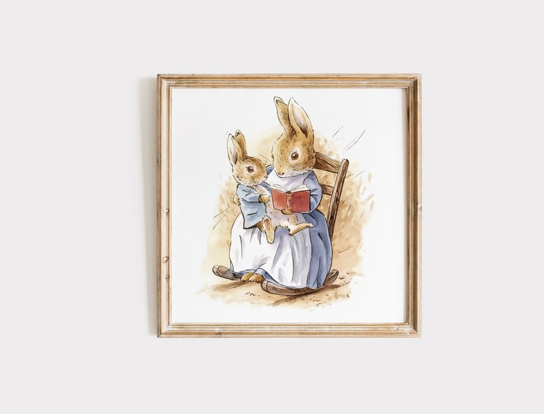 K&ouml;nnte beinhalten: Aquarellillustration von zwei Hasen. Einer, in einem blauen Kleid, liest einem kleineren Hasen in einer blauen Jacke aus einem Buch vor. Sie sitzen in einem Schaukelstuhl aus Holz. Das Kunstwerk ist in einem hellbraunen Holzrahmen gerahmt.
