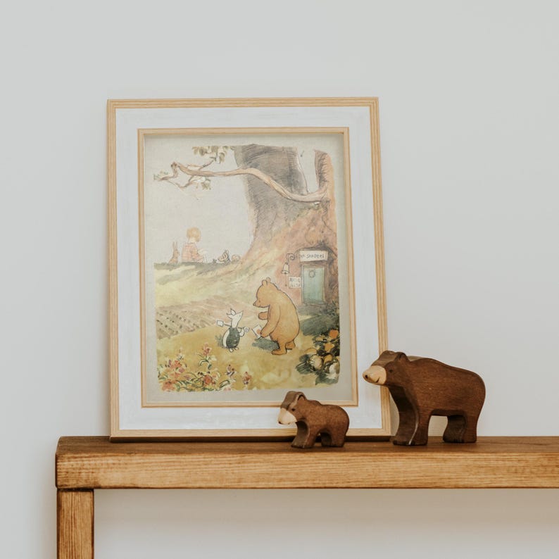 May include: A framed illustration of Winnie the Pooh and Piglet standing in front of a tree house. Two wooden bear figurines are in the foreground.