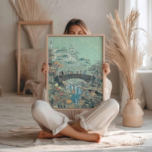May include: A framed art print featuring a fantasy landscape with a bridge, waterfall, and buildings. The artwork has a muted color palette with greens, blues, and browns. The frame is made of light-colored wood. The print is held by a person.