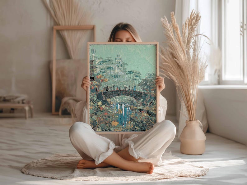 May include: A framed art print featuring a fantasy landscape with a bridge, waterfall, and buildings. The artwork has a muted color palette with greens, blues, and browns. The frame is made of light-colored wood. The print is held by a person.