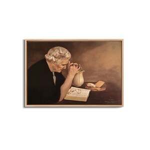 Op de afbeelding: Een ingelijste prent toont een oudere persoon met wit haar, handen gevouwen in gebed over een open boek. Een witte kan en brood liggen op een tafel. Het kunstwerk zit in een houten lijst.