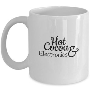 May include: A white ceramic mug with a curved handle. The mug features the text "Hot Cocoa & Electronics" in black script. The mug is designed for hot beverages.