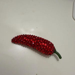 Puede incluir: Un objeto decorativo en forma de chile rojo cubierto de pequeñas gemas rojas brillantes. El tallo es verde. El objeto es probablemente un broche o un alfiler decorativo.