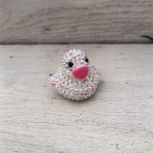 May include: A sparkly white duck ring with black eyes and a pink beak. The ring is made of rhinestones and is a fun and whimsical accessory.