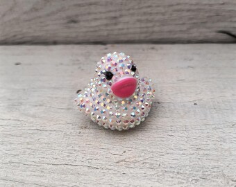 Puede incluir: Un anillo de pato blanco brillante con ojos negros y un pico rosa. El anillo está hecho de diamantes de imitación y es un accesorio divertido y caprichoso.