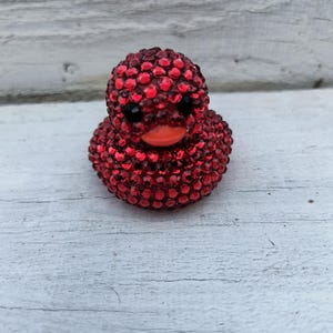 Puede incluir: Un pato decorativo rojo cubierto de brillantes pedrería. El pato tiene ojos negros y un pico naranja. El pato es un artículo de novedad, probablemente para exhibición o como pieza decorativa.