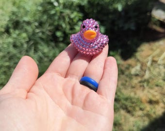 Puede incluir: Una pequeña figura de pato de goma completamente cubierta de brillantes diamantes de imitación de color rosa. El pato presenta un pico de color naranja brillante y dos pequeños ojos negros, creando una superficie texturizada y reflectante.