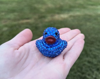 Puede incluir: Un pequeño pato de goma azul cubierto de brillantes piedras de cristal azules con un pico de plástico rojo.