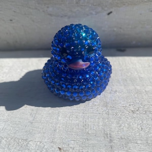 May include: A blue rubber duck covered in sparkling blue rhinestones. The duck has a pink beak and is sitting on a weathered, white surface. The duck is a decorative item.