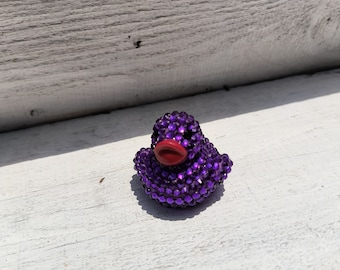 Puede incluir: Una pequeña figura de pato morado enjoyada con un pico rojo. El pato está cubierto de brillantes pedrería morada. La figura descansa sobre una superficie de madera blanca y desgastada.