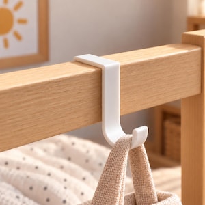 May include: A white, J-shaped over-the-door hook made of smooth plastic. The hook is securely mounted over the top edge of a light-colored wooden frame, with a beige fabric bag hanging from the bottom curve of the hook, providing storage.
