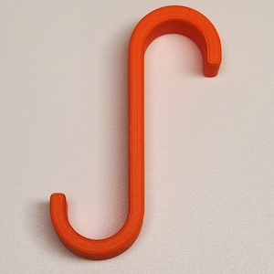 May include: An orange, S-shaped hook. The hook is made of a solid material and has a rounded shape on both ends. The hook is designed to hang items and is set against a plain, off-white background.