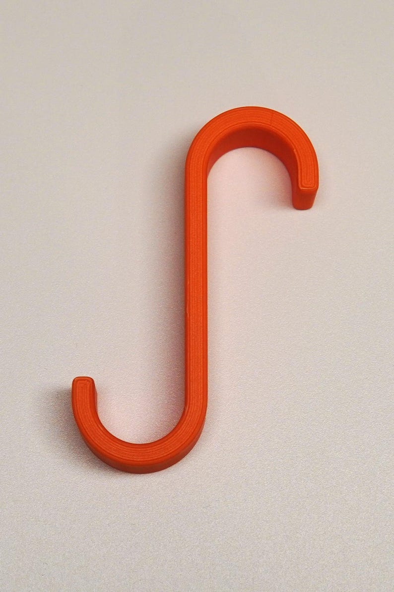 May include: An orange, S-shaped hook. The hook is made of a solid material and has a rounded shape on both ends. The hook is designed to hang items and is set against a plain, off-white background.